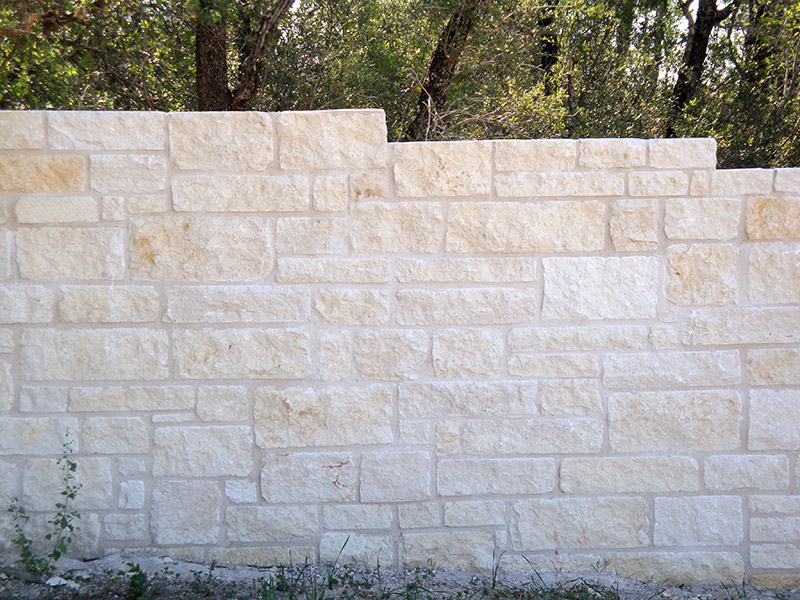 A light-colored stone wall with uneven top height, set outdoors with green trees and foliage visible in the background. Some small weeds grow at the base of the wall.