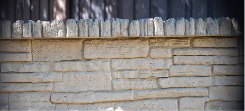 A beige stone wall with rectangular patterns is topped with irregular, vertically positioned stones. Shadows from nearby objects are visible on the upper portion of the wall.