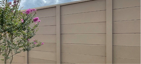 A beige concrete fence with horizontal panels stands behind a small tree with green leaves and clusters of pink flowers, under a partly cloudy sky.