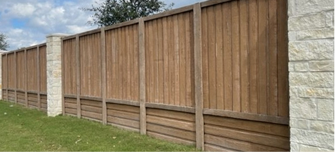 A wooden privacy fence with vertical panels and a stone pillar stands on a grassy lawn under a partly cloudy sky.