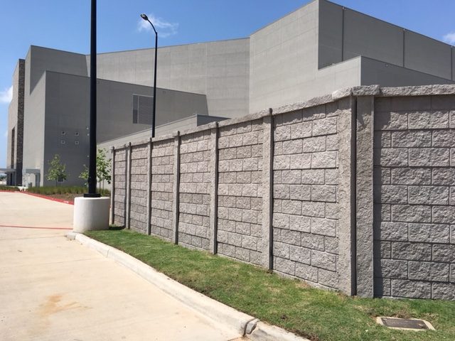A tall, gray concrete block wall runs alongside a sidewalk and grass strip, with a large, modern industrial or institutional building in the background and a streetlamp nearby under a blue sky.