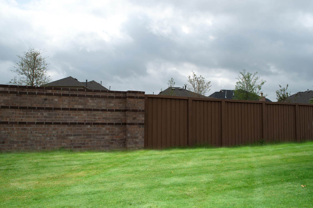 A tall brown wooden fence and a brick wall separate a grassy lawn from neighboring houses under a cloudy sky.