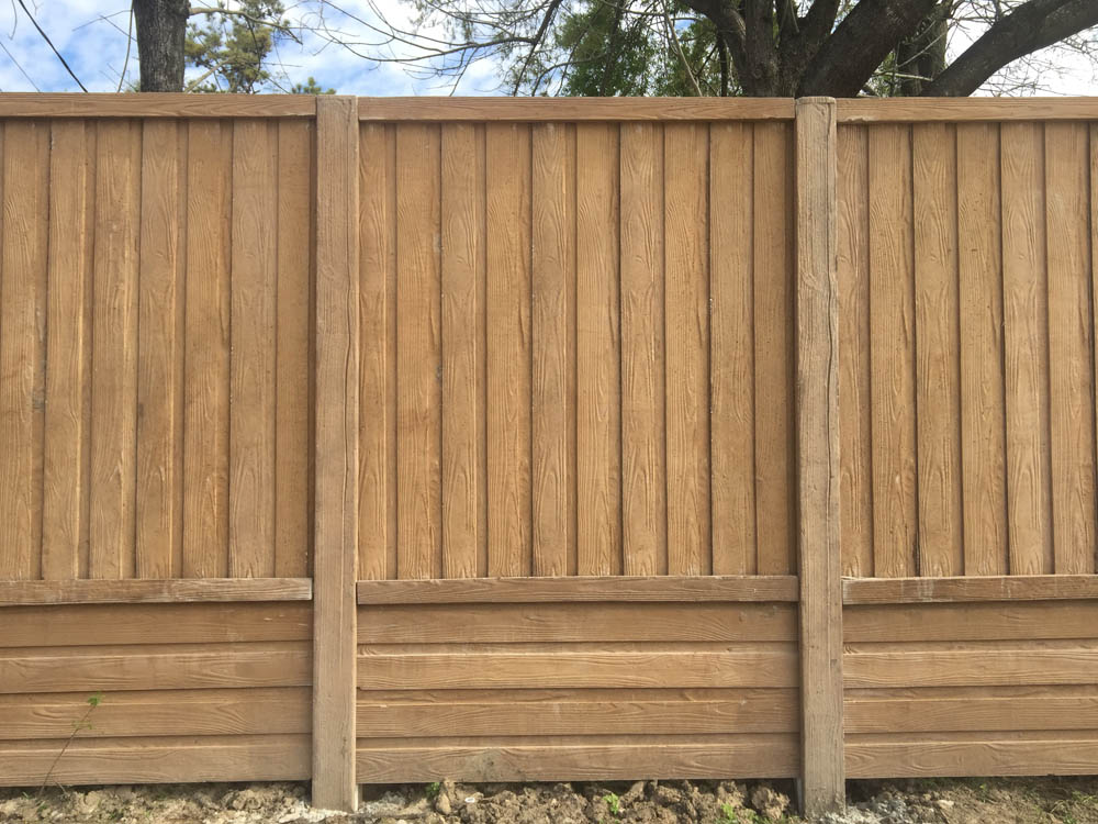 A tall wooden fence made of vertical and horizontal planks stands outdoors, with trees and a partly cloudy sky visible above the fence line. The ground at the base is bare soil.
