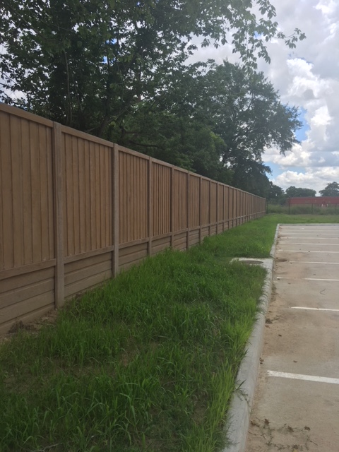 A long wooden privacy fence runs alongside a grassy area next to an empty concrete parking lot, with trees and a partly cloudy sky in the background.