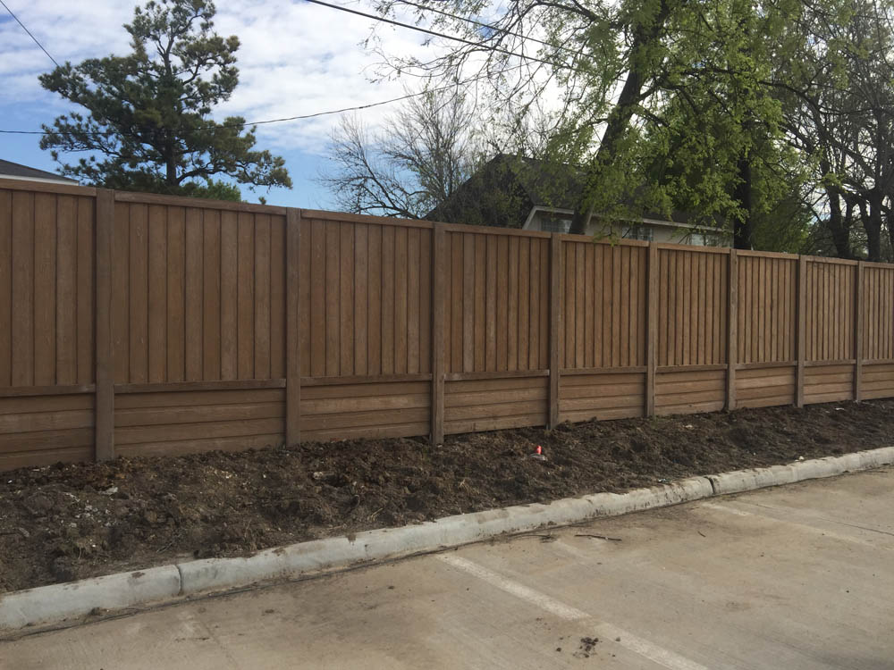 A tall wooden privacy fence runs alongside a paved road, with bare soil and some grass at its base. Trees and a house are visible behind the fence under a partly cloudy sky.