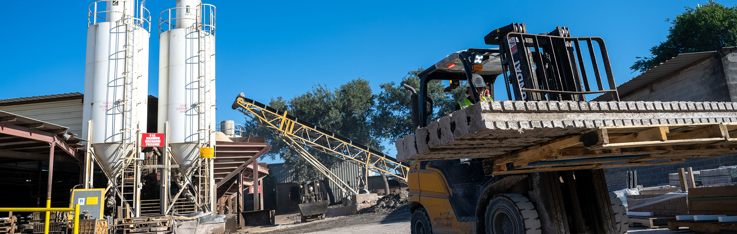 A forklift carries a stack of concrete blocks at an industrial site with tall silos, a conveyor belt, and various equipment under a clear blue sky.