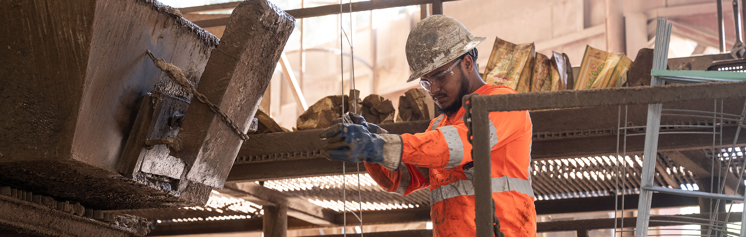 A construction worker in an orange safety jacket and helmet operates machinery in an industrial setting, handling materials with gloved hands amid a backdrop of metal beams and equipment.