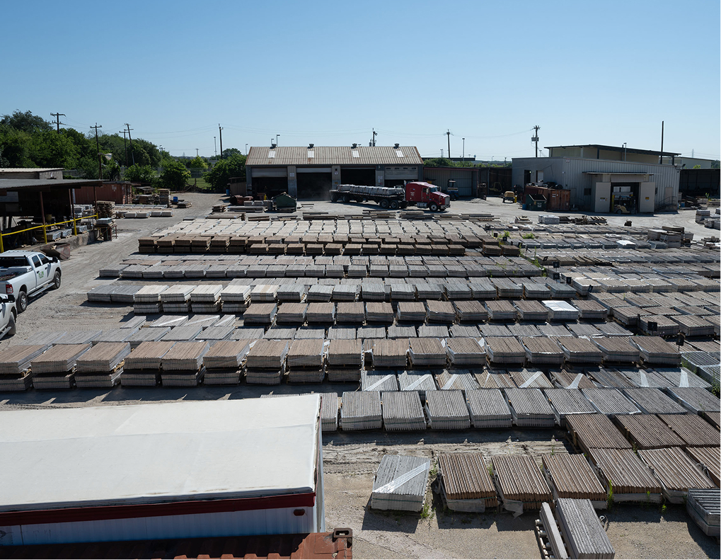 A large outdoor industrial yard with rows of stacked concrete slabs and blocks, trucks, and several warehouse buildings in the background under a clear blue sky.