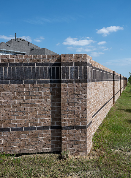 A tall brick wall with dark horizontal stripes extends alongside a grassy area under a blue sky with scattered clouds. The corner of a house roof is visible behind the wall.