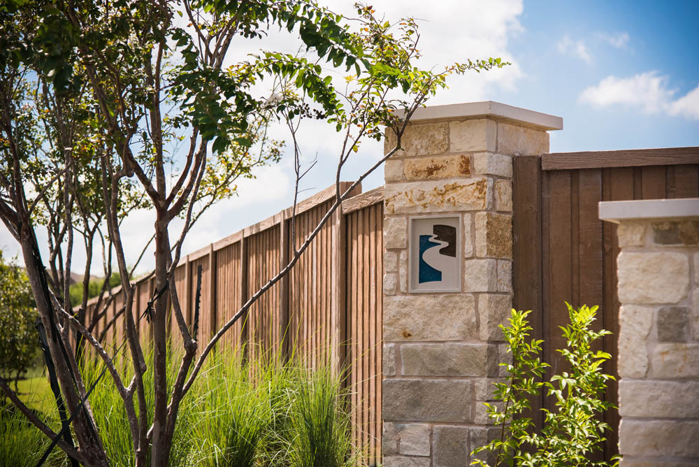 A stone pillar with a decorative tile stands next to a wooden fence, surrounded by green grass, shrubs, and a leafy tree under a blue sky with clouds.