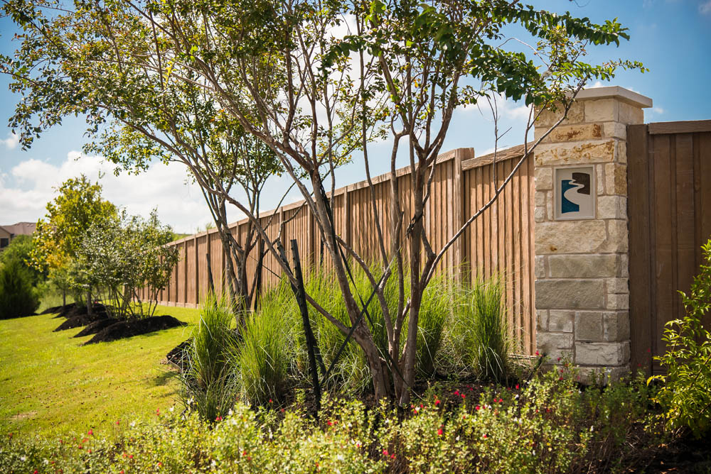 Landscaped area with trees, shrubs, and grass in front of a tall wooden fence with a stone pillar featuring a decorative plaque, under a bright, blue sky.