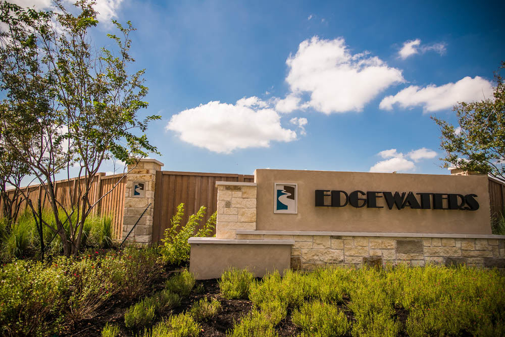 A stone entrance sign reads EDGEWATERS with a blue and white wave logo, surrounded by green shrubs and trees, under a partly cloudy blue sky.