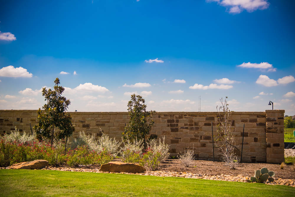 A stone wall with young trees and shrubs planted in front, bordered by neatly trimmed grass and rocks, under a bright blue sky with scattered clouds.