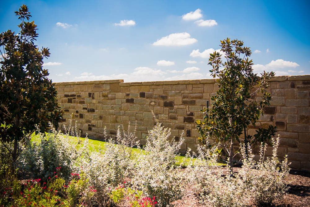 Landscaped garden with flowering shrubs and small trees in front of a tan stone wall under a bright blue sky with scattered clouds.