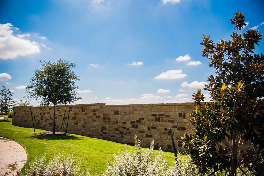 A stone wall runs across a green lawn under a bright blue sky with scattered clouds. Trees and shrubs are in the foreground, and sunlight casts shadows on the grass.