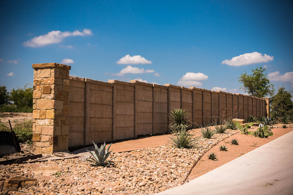 A stone and concrete wall runs alongside a landscaped area with rocks, desert plants, and agave under a bright blue sky with scattered clouds.