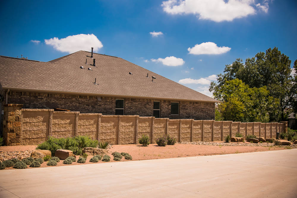 A tan stone privacy fence borders a backyard with landscaping of rocks and low plants, set in front of a brick house under a blue sky with scattered clouds.
