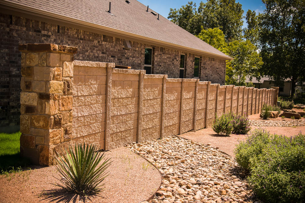 A tall stone and concrete privacy fence runs alongside a house with brick exterior walls, bordered by landscaped gravel, rocks, and various desert plants under a clear blue sky.