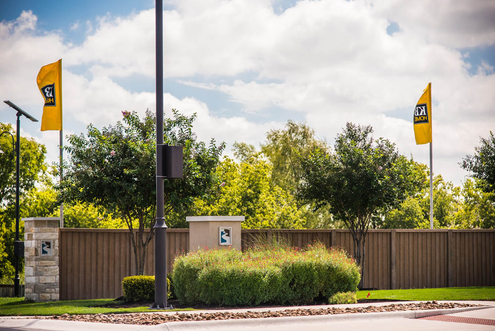A wooden fence with two yellow flags and a sign sits behind neatly trimmed bushes and trees, with a cloudy blue sky overhead and a street lamp in the foreground.