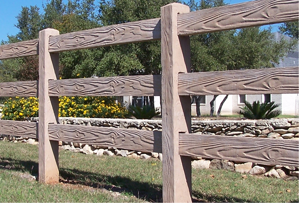 A close-up of a rustic wooden fence with three horizontal beams, set in a grassy yard. Yellow flowers, green bushes, stone edging, and trees are visible in the background.