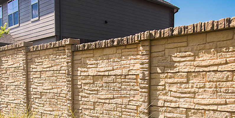 A beige stone-patterned privacy fence runs along the edge of a residential property, with a gray house and clear blue sky visible in the background.