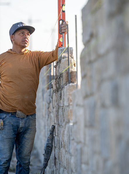 A construction worker in a brown hoodie and jeans uses a spirit level to check the alignment of a stone wall under construction, with yellow string lines guiding the placement of stones.