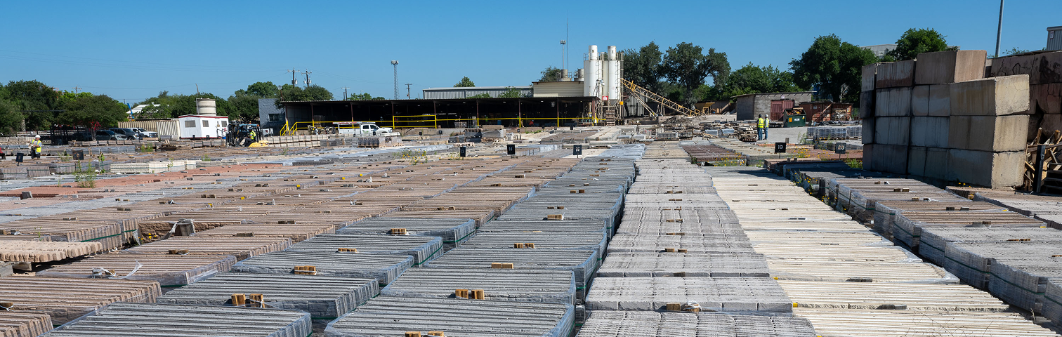 Rows of stacked stone slabs are neatly arranged in an outdoor storage yard under a clear blue sky, with industrial buildings, equipment, and some trees visible in the background.