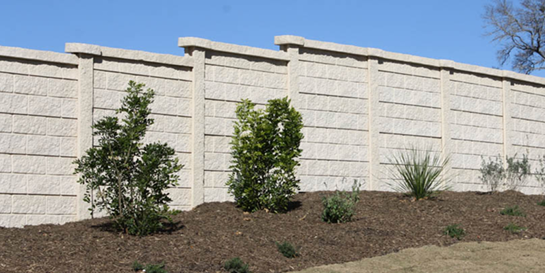 A beige concrete privacy wall with a textured surface stands behind a row of small green shrubs and plants in freshly mulched soil under a clear blue sky.