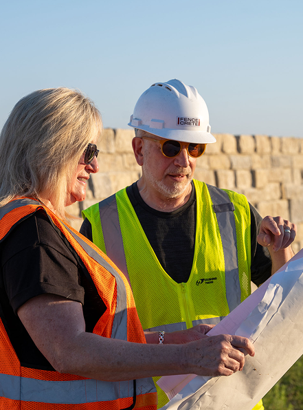 Two construction workers wearing safety vests and sunglasses, with one also wearing a hard hat, look over a large blueprint outdoors near a stone wall on a sunny day.
