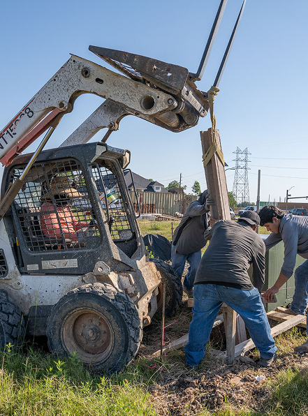 Three people work together to guide a wooden post being lifted by a small skid steer loader using a rope, on a grassy area near a road and houses under a clear blue sky.
