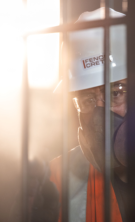 A construction worker wearing a white hard hat, safety glasses, orange vest, and a protective mask stands behind vertical metal bars in bright sunlight.