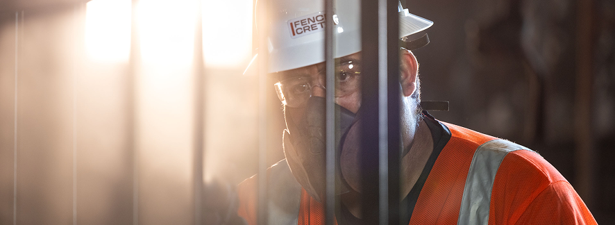 A construction worker wearing a hard hat, safety glasses, orange vest, and face mask works behind metal bars in a sunlit, dusty environment.