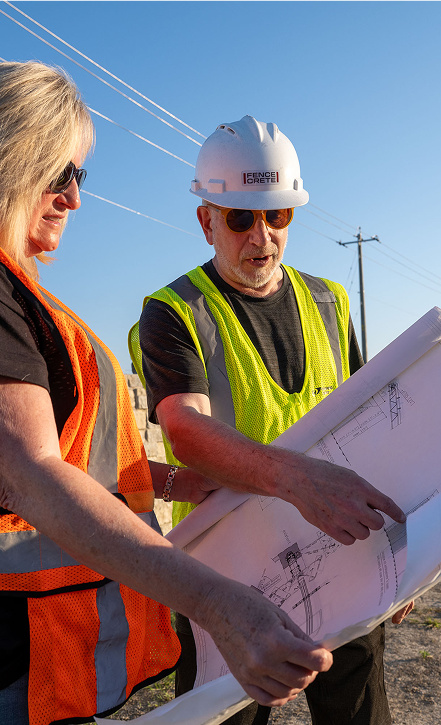 Two construction workers wearing safety vests and hard hats examine blueprints outdoors on a sunny day. One points to the plans while the other holds the paper, with power lines and a clear blue sky in the background.
