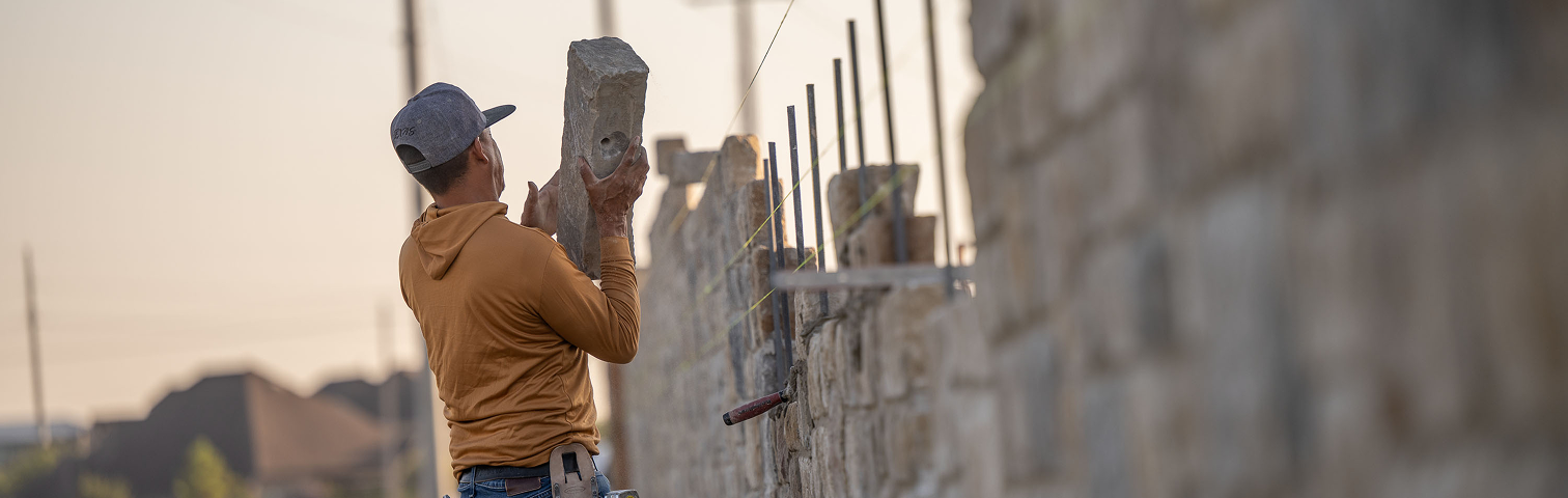 A construction worker wearing a brown hoodie and gray cap lifts a large stone to build a stone wall outdoors. Metal rods and construction tools are visible nearby under a clear sky.