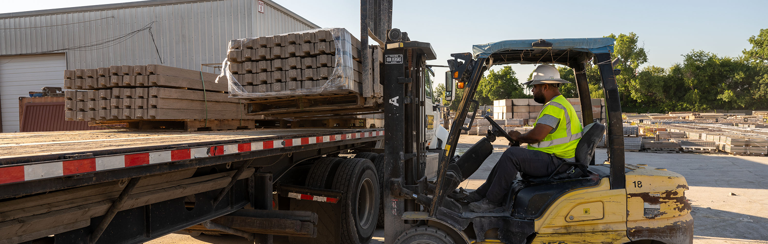A worker in a safety vest and helmet operates a forklift to load stacked metal pipes onto a flatbed truck in an outdoor industrial yard.