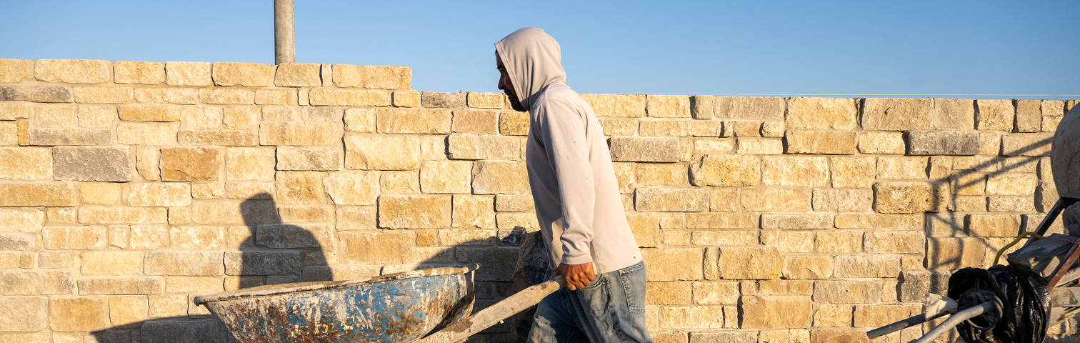 A person in a hoodie pushes a worn blue wheelbarrow past a stone wall under a clear sky, with long shadows cast by the sunlight.