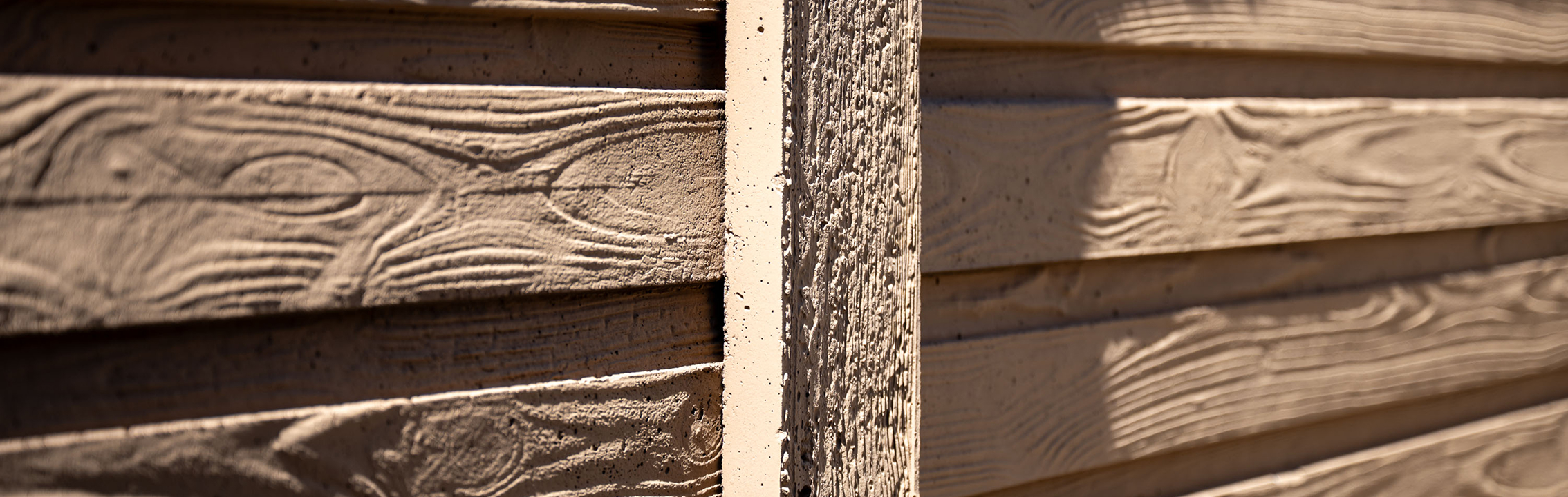 Close-up of tan-colored wooden siding with a textured, horizontal pattern. Sunlight creates shadows, highlighting the wood grain and the vertical beam dividing the panels.