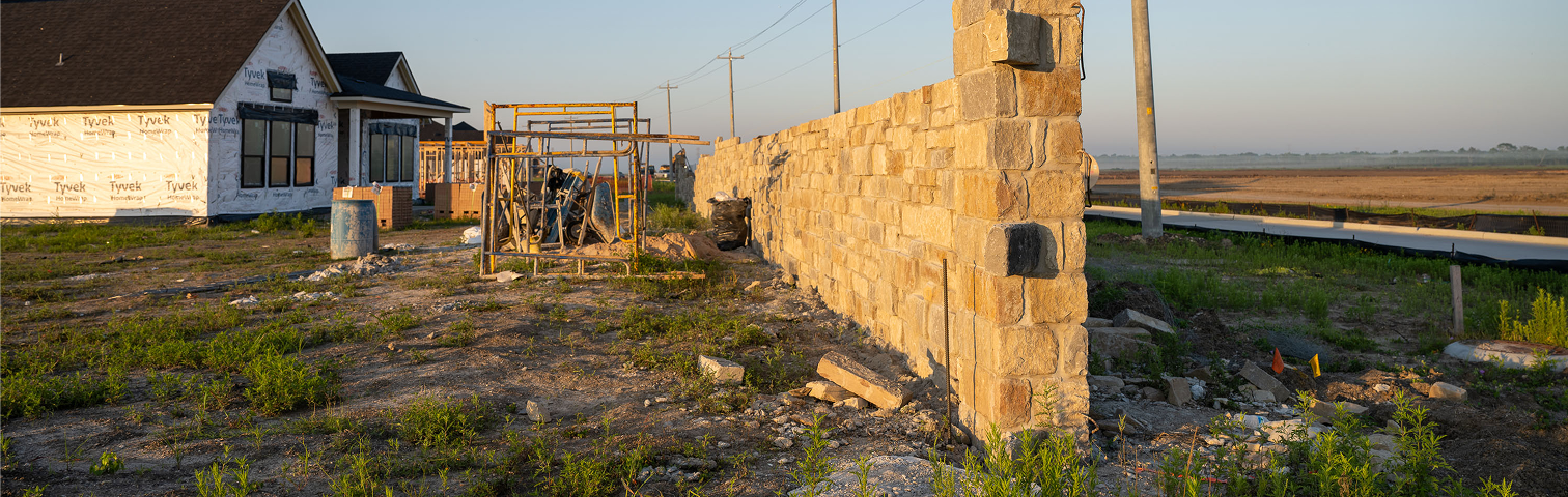 A partially constructed stone wall stands on a dirt lot near a house under construction, with building materials scattered around and fields visible in the background under a clear sky.