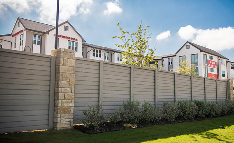 A tall, beige privacy fence with stone pillars stands in front of modern apartment buildings with Now Leasing banners, green shrubs, and a blue sky with scattered clouds.