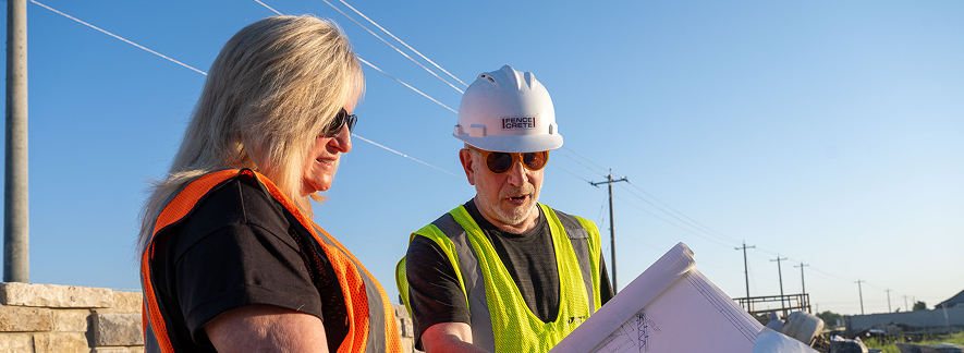 Two construction workers wearing safety vests and hard hats review blueprints outdoors near power lines and a stone wall on a sunny day.