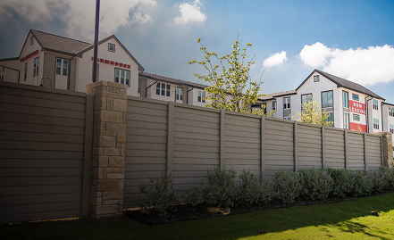 A row of modern apartment buildings with Now Leasing signs is visible behind a tall gray privacy fence, with some shrubs and a small tree in the foreground under a partly cloudy sky.