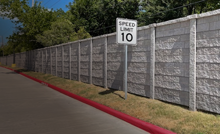A Speed Limit 10 sign stands beside a concrete wall, with a red-painted curb and a paved road on the left, under a partly cloudy sky with trees in the background.