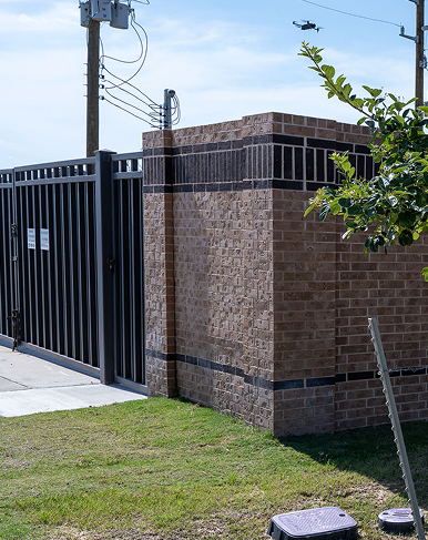A black metal gate and brick wall on a grassy area, with overhead power lines, a utility pole, and a small tree branch visible. The sky is clear and blue.