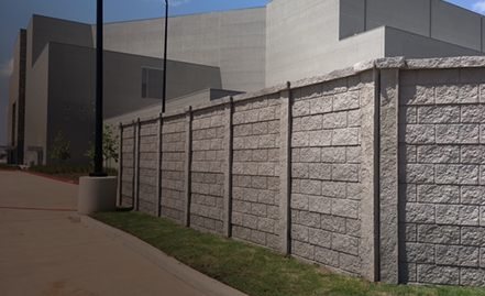 A tall concrete wall with a textured block pattern runs along a sidewalk, partially enclosing a modern, windowless building in the background under a clear sky.