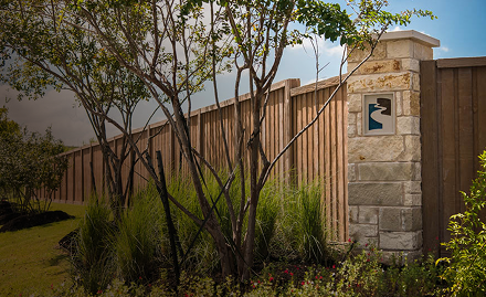 Tall trees and green shrubs grow in front of a long wooden fence with a stone pillar that features a small decorative cutout. The scene is outdoors with partly cloudy skies.