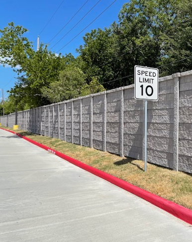 A concrete road beside a tall gray brick wall with a Speed Limit 10 sign. Green trees rise behind the wall under a clear blue sky, and the curb along the road is painted red.