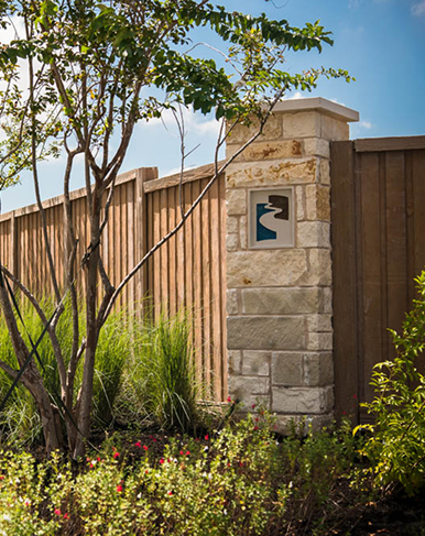 A stone pillar with a decorative inset is set along a wooden fence, with landscaping that includes green grass, bushes, and a tree under a partly cloudy sky.