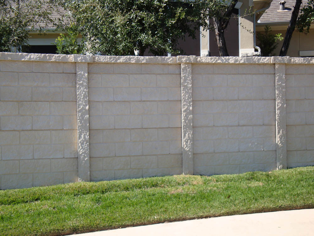 A beige stone privacy wall with vertical columns separates a grassy lawn from a house and trees in the background on a sunny day.