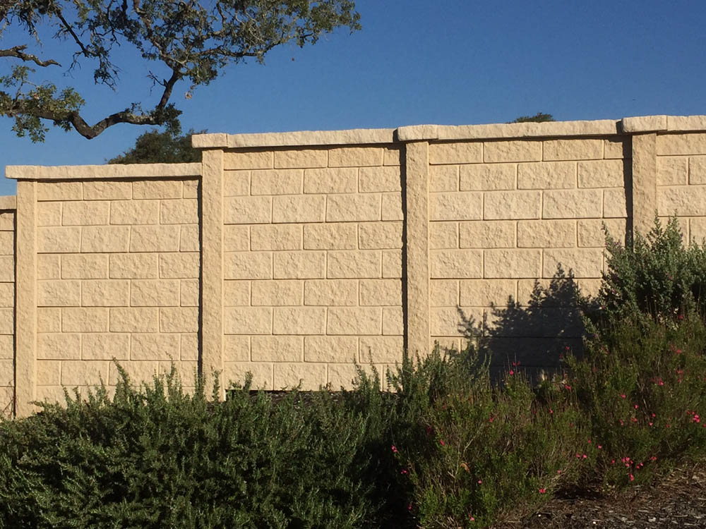 A beige stone-patterned concrete wall stands under a clear blue sky, with green shrubs and small pink flowers growing at its base. A tree branch extends into the frame from the upper left corner.