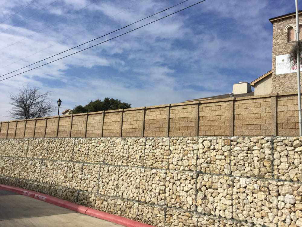 A tall wall made of stacked light-colored stones on the bottom and tan concrete panels on top runs alongside a street curb under a partly cloudy sky. A building and trees are visible in the background.